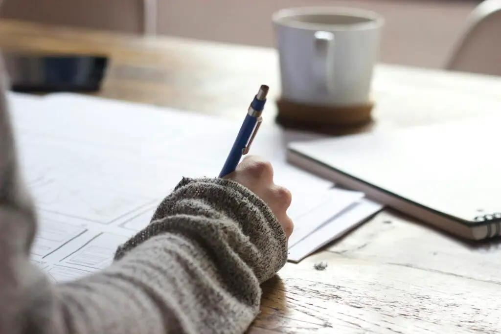 hand holding a pen writing on papers on a desk. Coffee cup sitting on desk in the background.